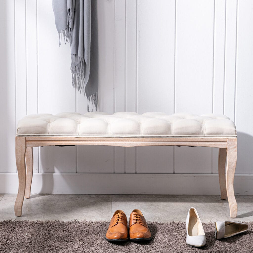 Beige tufted entryway bench with carved wood legs placed by a wall with shoes in front
